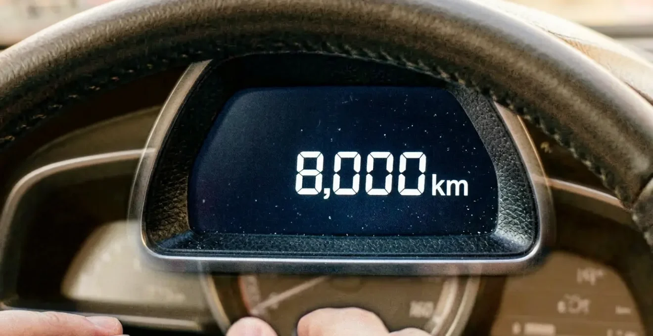 Gros plan sur un compteur kilométrique moderne affichant 8000 km avec des mains sur le volant et un paysage urbain flou en arrière-plan